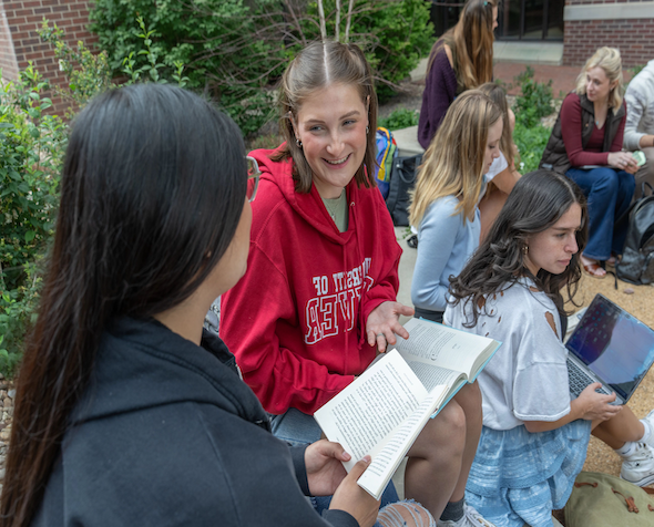 A student in a University of Denver sweatshirt holds open a book while talking with another student on the University of Denver campus. 